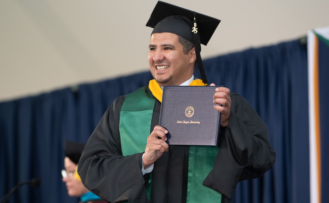 Male graduate student proudly holding his diploma on stage at commencement and smiling at the crowd