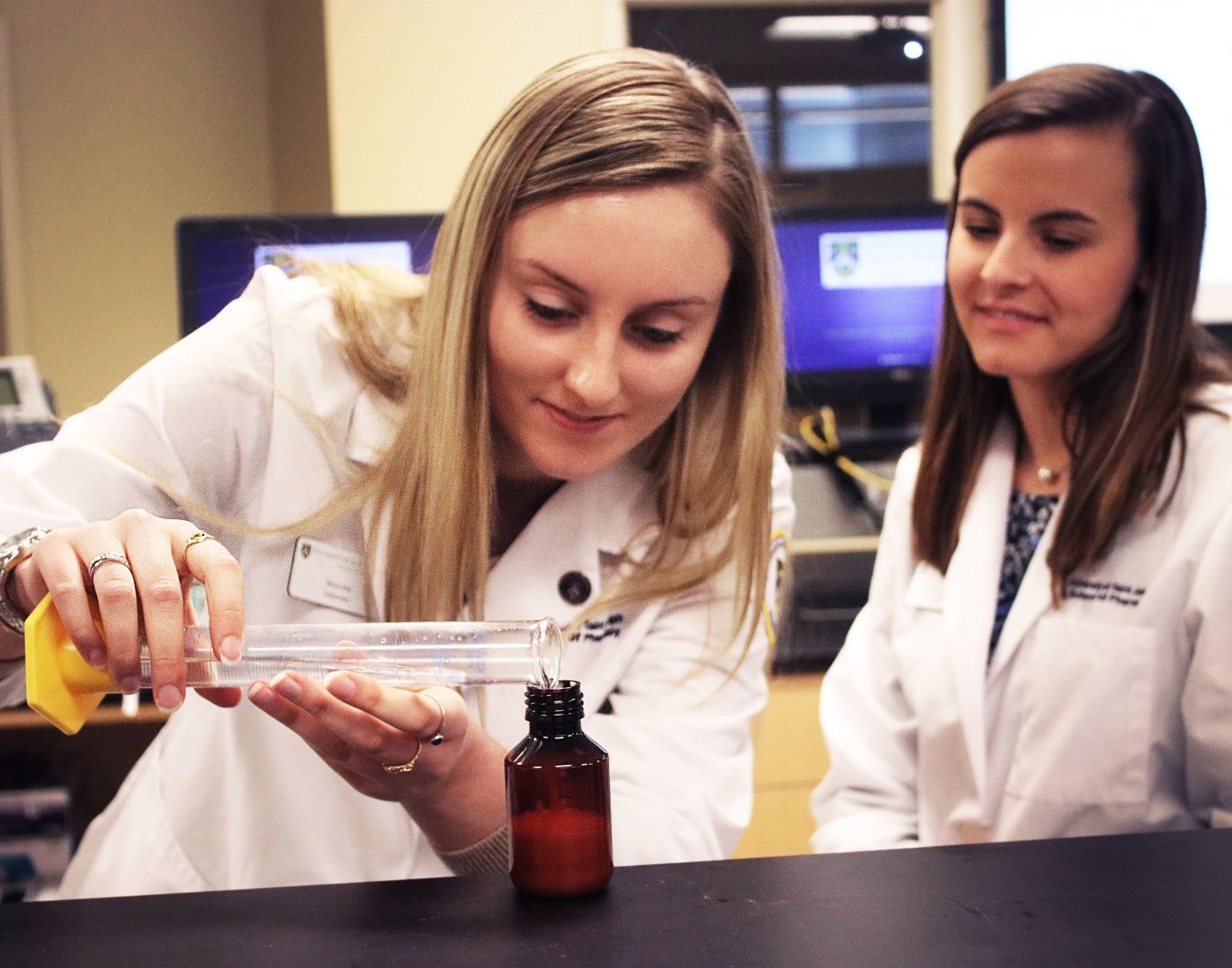 two students in lab coats pouring liquid from beaker