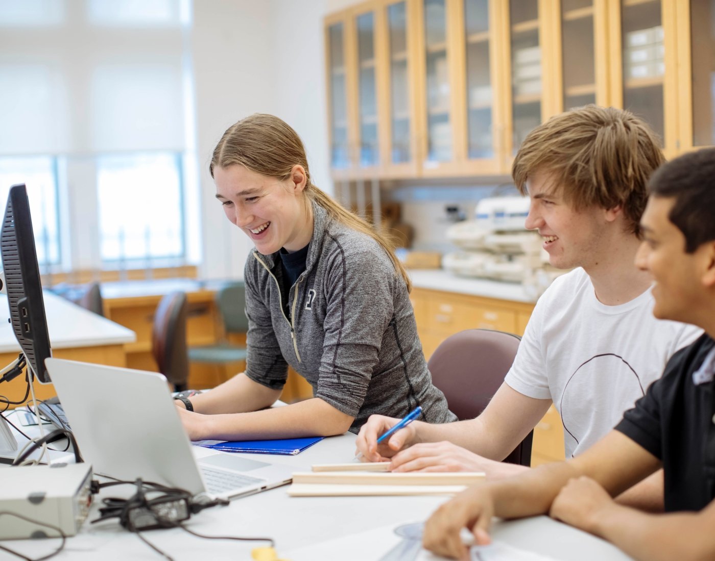 three students sitting a lab table with computer screens in front of them, with happy expressions