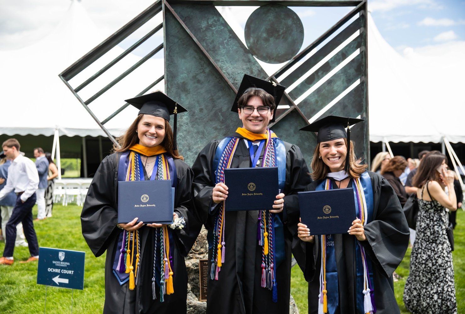 Three students smiling and holding their diplomas in their cap and gowns outside.