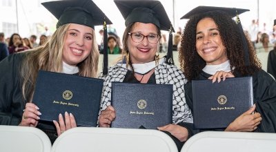 Three graduate students sitting and smiling with their diplomas in hand, in their caps and gowns.