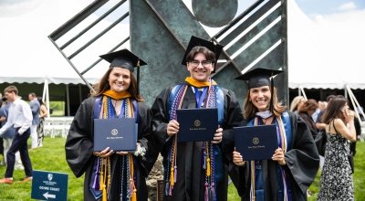 Three students smiling and holding their diplomas in their cap and gowns outside.