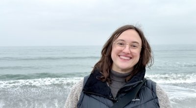 Photo of Brennan at the beach, smiling in a turtleneck and vest.